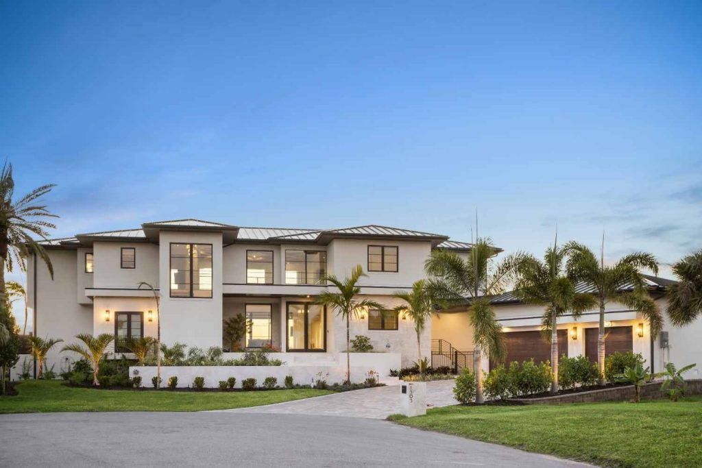 White modern waterfront mansion at dusk with palms and lit interiors